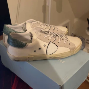 Vita sneakers med gröna detaljer från philipe model - HELT NYA Snygga vita sneakers från Philippe Model med gröna detaljer på hälen. Skorna har snörning med text och en unik design på sidan. Perfekta för en stilren look. Strlk 42, box tillkommer. Skriv ifall i frågor.