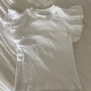 Vit ribbad topp med volangärmar - Säljer en söt vit ribbad topp med volangdetaljer på ärmarna. Perfekt för en stilren och chic look. Toppen har en rund halsringning och är tillverkad i ett mjukt material.
