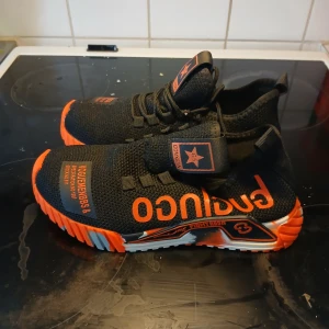 Svarta och orange sneakers med textdetaljer - Snygga svarta sneakers med orange detaljer och text på ovansidan. Skorna har en unik sula med ett färgglatt mönster i orange, svart och vitt. De är utrustade med snörning och har en modern design som sticker ut.