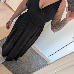 Svart omlottklänning - Elegant svart omlottklänning med v-ringning och knytbara axelband. Klänningen har en smickrande passform med smockdetaljer i midjan som ger en fin siluett. Perfekt för en stilren look.