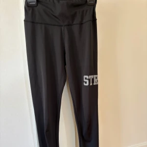 Svarta leggings med text från OTP - Snygga svarta leggings med hög midja och texten 'STRONG' i silver på benet. Perfekta för träning eller en avslappnad dag. Gjorda i ett stretchigt syntetmaterial för bästa komfort.