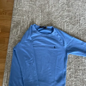 Blå tröja från Polo Ralph Lauren - Säljer en snygg blå tröja från Polo Ralph Lauren. Tröjan är tillverkad i 100% bomull och har en broderad logotyp på bröstet. Perfekt för en stilren och avslappnad look.