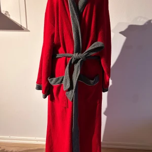 Röd morgonrock med grå detaljer - Säljer en mysig röd morgonrock från Hemtex. Den har en huva, fickor och ett knytbälte i midjan för en bekväm passform. Perfekt för att hålla sig varm och bekväm hemma.  