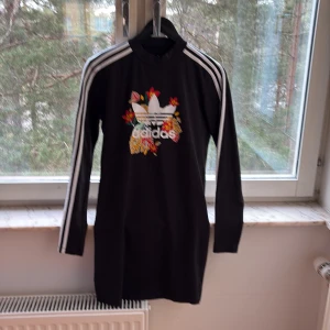 Svart klänning från Adidas - Snygg svart klänning från Adidas med blommigt tryck och klassiska vita ränder längs ärmarna. Klänningen har långa ärmar och en normal passform. Perfekt för en sportig och trendig look.