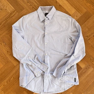 Ljusblå skjorta från Armani Exchange - Säljer en stilren ljusblå skjorta från Armani Exchange i storlek M. Skjortan har en klassisk krage och knappar framtill. Den är broderad med A|X-loggan på bröstet och har långa ärmar med knappslå vid ärmsluten. Slim fit. Liten fläck bak och lite solblekt i kragviket