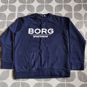 Mörkblå tröja från Björn Borg - Säljer en stilren mörkblå tröja från Björn Borg med vit text "BORG SPORTSWEAR" på framsidan. Tröjan har långa ärmar och en klassisk rund halsringning. Perfekt för en avslappnad stil.