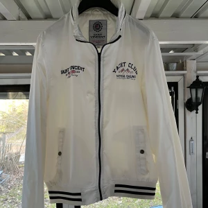 Vit vindjacka från Saint Vincent - Snygg vit vindjacka från Saint Vincent med schyssta detaljer på bröstet. Jackan har dragkedja och ribbade muddar i mörkblått och vitt. Finns även en luva gömd i kragen med dragkedja. Perfekt för en stilren look. Storlek L men passar M också.