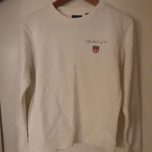 Vit tröja från GANT - Säljer en stilren vit tröja från GANT med deras logotyp och emblem på bröstet. Tröjan har långa ärmar och en rund halsringning. 
