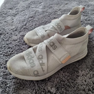 Beige sneakers från Adidas - Säljer ett par sneakers från Adidas med elastiska band över foten. Med lite omsorg och en tvätt blir dom nog som nya.