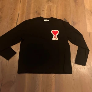 Svart tröja från Ami Alexandre Mattiussi - Snygg svart tröja från Ami Alexandre Mattiussi med ett rött hjärta på bröstet. Tröjan har långa ärmar och en klassisk passform. Perfekt för en stilren look.