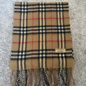 Rutig halsduk från Burberry - Säljer en klassisk rutig halsduk från Burberry i beige, svart och röd. Den har fransar i ändarna och är perfekt för att hålla dig varm och stilren under kyliga dagar.