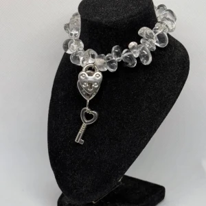 Armband med hjärtformad låsdetalj - Unikt armband med klara pärlor och en hjärtformad låsdetalj i silver. Armbandet har en charmig nyckel som hänger från låset, vilket ger en lekfull och romantisk touch. Perfekt för att ge en personlig stil till din outfit.