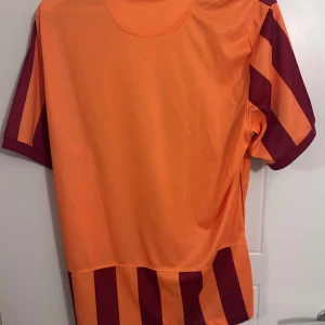 23-24 Galatsray forbollströja - Säljer en randig fotbollströja från Nike i orange och vinrött. Tröjan har korta ärmar och en klassisk krage. Perfekt för fotbollsfans som vill visa sitt stöd med stil.