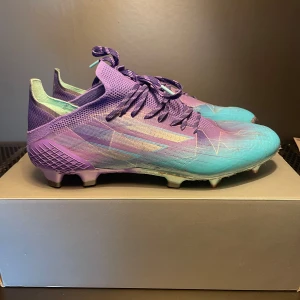 Adidas fotbollsskor i lila och turkos - Snygga fotbollsskor från Nike i en cool lila och turkos färgkombination. Skorna har snörning och en modern design med mönstrade detaljer. Perfekta för att dominera på planen med stil.