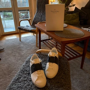 Valentino Open sneakers i vitt och svart - Snygga Valentino Open sneakers i vitt med en bred svart rem över ovansidan. Skorna har en klassisk design med snörning och en unik detalj av små nitar runt sulan. Perfekta för en stilren look. inga skador med få tecken på användning perfekt sick och sällan använda