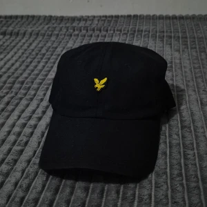 Svart keps från Lyle & Scott - Snygg svart keps från Lyle & Scott med en gul broderad logotyp framtill. Kepsen har justerbar rem baktill för perfekt passform. Perfekt för en casual look.