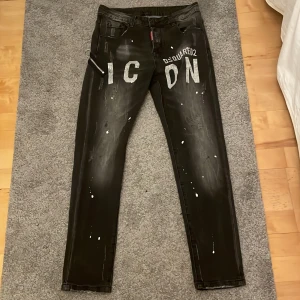 Svarta jeans med tryck från Dsquared2 - Snygga svarta jeans från Dsquared2 med coolt tryck 'ICON' i vitt på framsidan. De har en unik design med slitningar och vita färgstänk för en edgy look. Dragkedjedetalj med ring på sidan ger extra stilpoäng. Perfekta för dig som vill sticka ut!