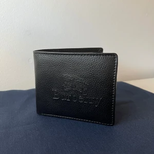 Svart plånbok från Burberry - Snygg svart plånbok från Burberry i skinn med präglad logotyp på framsidan. Plånboken har flera kortfack och en knappstängd myntficka. Perfekt för att hålla ordning på dina kort och kontanter✅ Säljer denna plånbok för 699kr ✅👏