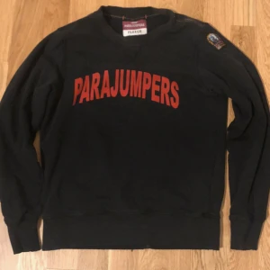 Svart fleece tröja från Parajumpers - Säljer en svart fleece tröja från Parajumpers med röd text på framsidan. Tröjan har långa ärmar och en broderad patch på ärmen. Perfekt för kyliga dagar.