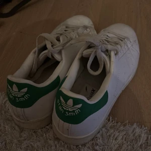 Vita Adidas Stan Smith sneakers  - Säljer ett par klassiska vita Adidas Stan Smith sneakers med gröna detaljer. Skorna har snörning och rund tå, perfekt för en stilren look. (Fejk, men identiska till riktiga Adidas skor) 