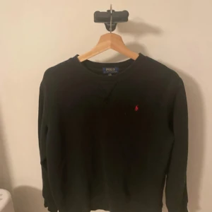 Svart tröja från Polo Ralph Lauren - Säljer en klassisk svart tröja från Polo Ralph Lauren med det ikoniska röda broderade logotypen på bröstet. Tröjan har långa ärmar och en rund halsringning. Perfekt för en stilren och avslappnad look. Lite små fläckar på mudrarna men för priset så finns det inget att klaga på😁 pris kan diskuteras 