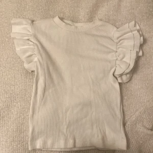 Super fin vit ribbad topp från Gina Tricot - Säljer en söt vit ribbad topp från Gina Tricot med volangdetaljer på ärmarna. Perfekt för en sommar eller vår dag.  Passar bra till både jeans, shorts och kjol. 🌟nyt skick, aldrig använd men lagen är tyvärr borta. Inga fläckar. Inga hål.