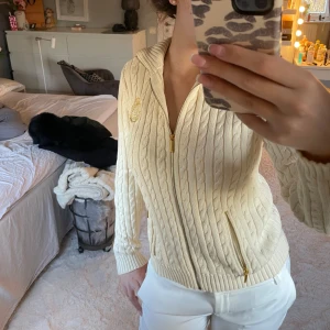 Beige stickad kofta från Ralph Lauren - Säljer en elegant beige/krämvit stickad kofta från Ralph Lauren med kabelstickat mönster och dragkedja framtill. Koftan har en broderad logga på bröstet och långa ärmar. Perfekt för en stilren look.