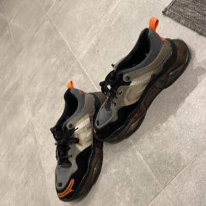 Svarta och grå sneakers med orange detaljer - Snygga sneakers i svart och grått med coola orange detaljer och texten 'WHAT DO YOU WANT?' på framsidan. Skorna har en robust sula och snörning för en säker passform. Perfekta för en trendig look.