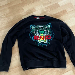 Svart Kenzo sweatshirt med tigertryck - Svart sweatshirt från Kenzo med ikoniskt broderat tigerhuvud i grönt, blått och gult på bröstet samt röd Kenzo-text. Rund hals och ribbade muddar.