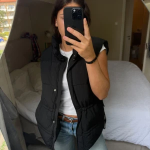 Svart quiltad dunväst med hög krage - Svart dunväst med hög krage och quiltad design. Västen har dragsko i midjan för snygg passform och stängs med både dragkedja och tryckknappar. Perfekt att slänga över en hoodie eller t-shirt för en chill look.