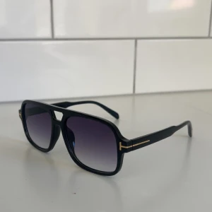 Svarta Tom Ford solglasögon - Snygga svarta solglasögon från Tom Ford med fyrkantig form och coola gulddetaljer på skalmarna. Glasen är tonade i mörkgrått och bågen är tillverkad i blank syntet. Perfekt för dig som vill ha en stilren och modern look.