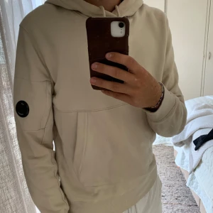 Beige hoodie från C.P. Company - Säljer en beige hoodie från C.P. Company med klassisk huva och snörning. Tröjan har en stor ficka framtill och den ikoniska svarta linsen på vänster ärm.  Den är i väldigt fint skicka och är självklart äkta✅