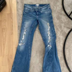 Blå bootcut jeans med slitningar - Säljer ett par blå jeans med bootcut och snygga slitningar framtill. Jeansen har låg midja, klassiska fem fickor och coola detaljer på bakfickorna med knappar. Perfekt för dig som gillar en avslappnad och trendig look.