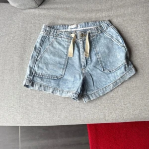 Blå jeansshorts med snörning - Snygga ljusblå jeansshorts från H&M med snörning i midjan och stora fickor fram. Modellen är loose fit och har en bakficka. Perfekta för sommaren och chill dagar. Klassisk knapp och dragkedja framtill.