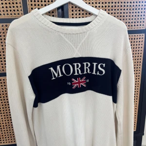 Vit stickad tröja Morris - Snygg vit stickad tröja från Morris med bred marinblå rand över bröstet och broderad logga samt brittisk flagga. Tröjan har rund hals och långa ärmar, perfekt för dig som gillar klassisk stil med en modern twist.