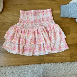 Blommig volangkjol från RVLS - Supersöt kort kjol från RVLS med volanger och blommigt mönster i rosa och orange toner på vit botten. Kjolen har smockad midja som ger en skön och flexibel passform. Perfekt för dig som gillar färg och lekfull stil!