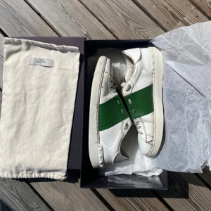 Valentino open - Snygga vita sneakers från Valentino Garavani med en bred grön läderdetalj över sidan. Skorna har rund tå, platt sula och klassisk snörning. Kommer med original dustbag och låda. Perfekt för dig som gillar exklusiva och stilrena sneakers. Liten defekt på högerskott där man ser en spricka men inget som förstör skorna eller annat. Skick 8/10