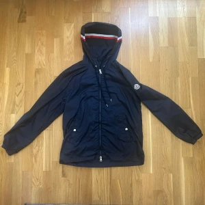 Mörkblå vindjacka från Moncler - Snygg mörkblå vindjacka från Moncler med huva och dragkedja framtill. Jackan har vita och röda detaljer på huvan, två fickor med knappar och klassisk Moncler-logga på ärmen. Perfekt för dig som vill ha en stilren och sportig look.