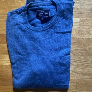 Blå stickad tröja från Lexington - Snygg blå stickad tröja från Lexington i storlek S. Tröjan har rund hals och en enkel, klassisk design med en liten ficka framtill. Perfekt för dig som gillar stilrena och bekväma plagg med lite preppy känsla.