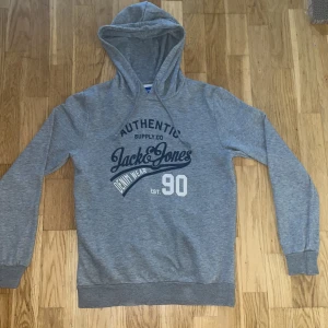 Grå hoodie från Jack & Jones storlek S - Säljer en grå hoodie från Jack & Jones med tryck i mörkblått och vitt på bröstet. Tröjan har huva med snörning och långa ärmar, samt ribbade muddar vid ärmslut och nederkant. Perfekt för chill dagar och streetstyle. Tröjan är i storlek S. Tröjan är i jätte bra skick och ifall ni har några funderingar kom dm.