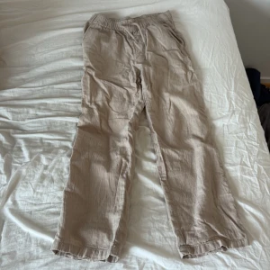 Beiga linnebyxor från Kappahl - Snygga beiga linnebyxor från Kappahl med avslappnad passform och resår i midjan. Byxorna har raka ben och sidfickor, perfekta för en chill och luftig stil. Materialet är linne som ger en skön känsla, och färgen är lätt att matcha med det mesta.