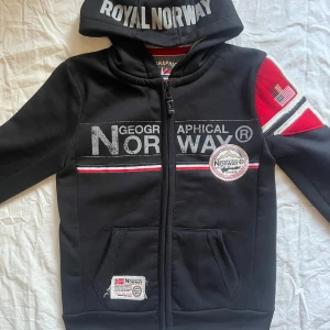 Svart hoodiejacka Geographical Norway - Svart hoodiejacka från Geographical Norway med dragkedja, stora tryck och broderade märken framtill och på ärmen. Jackan har huva med tryck, röda och vita detaljer samt fickor framtill. Perfekt för dig som gillar sportig och street-inspirerad stil.