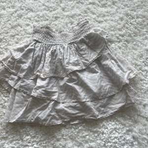 Randig volangkjol från H&M - Supersöt kortkjol från H&M med ljusgrå och vita ränder. Kjolen har bred smock i midjan och dubbla volanger som ger en flowy och lekfull look. Perfekt för dig som gillar mjuka material och sköna passformer.