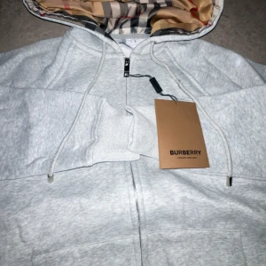 Grå hoodie från Burberry med mönstrad huva - Snygg ljusgrå hoodie från Burberry i bomull med dragkedja och klassisk rutmönstrad huva i silkesliknande material. Tröjan har snörning i huvan, ribbade muddar och känguruficka framtill. Perfekt för dig som vill ha en stilren men ändå lyxig look.