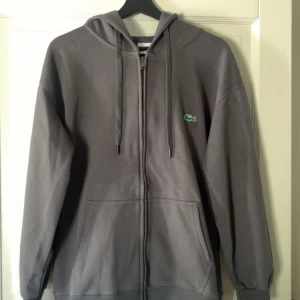 Grå hoodie från Lacoste med dragkedja - Snygg grå hoodie från Lacoste med klassisk krokodillogga på bröstet. Tröjan har huva med snörning, hel dragkedja och två stora fickor framtill. Regular fit och tillverkad i mjuk bomull, perfekt för chill dagar.