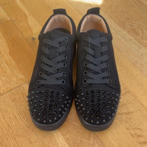 Christian Louboutin svarta sneakers med nitar - Svarta sneakers i mocka från Christian Louboutin med coola svarta nitar över tån och den ikoniska röda sulan. Skorna har snörning och en rund tå, perfekt för dig som vill sticka ut med en edgy vibe.