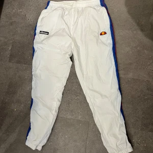 Vita trackpants från Ellesse L - Snygga vita trackpants från Ellesse med blå och röda detaljer längs sidorna. Byxorna har resår i midjan, snörning och muddar vid bensluten. Klassisk Ellesse-logga på ena benet och patch på andra. Perfekta för en sportig och retro look.