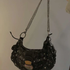 Svart axelväska från Guess med kedja - Cool svart axelväska från Guess med silverfärgade detaljer, kedjerem och logomönster över hela väskan. Väskan har nitar, metallspänne framtill och en stor G-charm. Perfekt storlek för det viktigaste och ger en edgy vibe till din outfit.                                #GuessBag #GuessVäska #DesignerVäska #Axelremsväska #Kedjerem #Modefynd #PrelovedFashion #SecondHandVäska #Y2KStyle #VintageGuess #Streetstyle #BrandedBag #GuessLovers #Modeinspo #FashionFinds