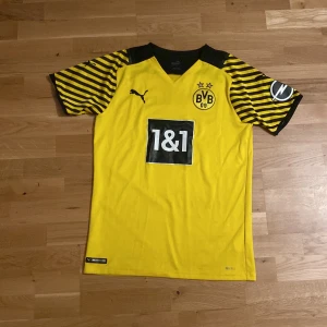 Borussia Dortmund matchtröja Puma S - Borussia Dortmunds officiella matchtröja från säsongen 2021/2022 gjord av Puma i storlek S. Tröjan är gul med svarta detaljer och har svarta och gula ränder på ärmarna. BVB-logga och sponsortryck på bröstet, samt DORTMUND-tryck på ryggen. Tillverkad i lätt och ventilerande polyester. Har en liten fläck längst ner vid tröjan som inte märks av alls.