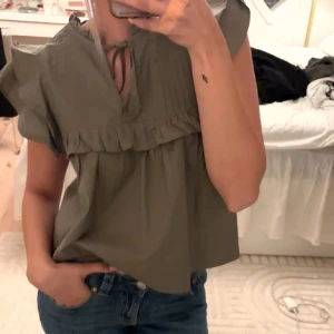 Olivgrön blus med volang och knyt - Supersöt olivgrön blus med volangdetaljer och knyt vid halsen. Luftig modell med korta ärmar och lätt rynk under bysten. Perfekt till jeans för en chill men ändå stylish look. Lånade bilder och priset kan diskuteras. Aldrig använd säljer då den var för liten.